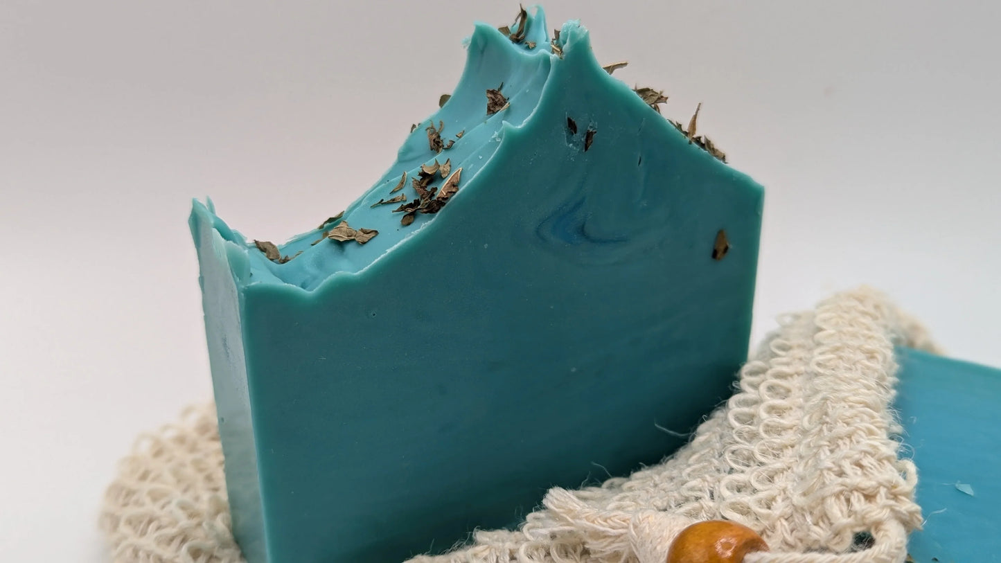 Blue handmade soap bar with dried herbs on top, resting on a natural soap saver pouch