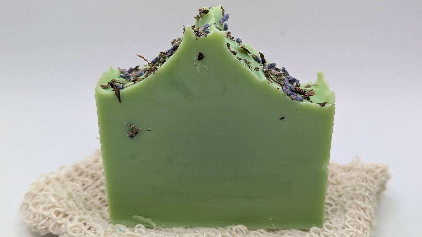 green handmade soap bar with dried herbs and flowers on top, placed on a natural soap saver